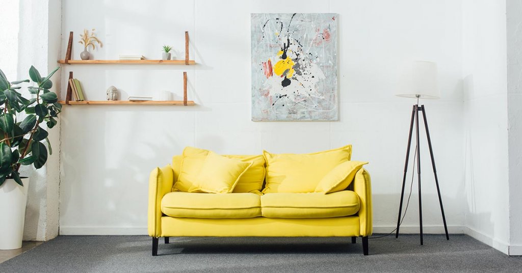 Ein Zimmer mit einem gelben Sofa, einer Lampe, Pflanzen sowie einem Bild und einem Regal an der Wand | Home Staging