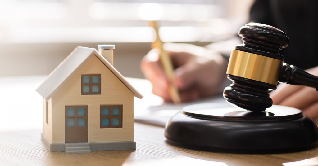 Ein Mann unterschreibt ein Dokument auf einem Tisch und im Vordergrund stehen ein Modellhaus und ein Richterhammer | Immobilie in der Scheidung